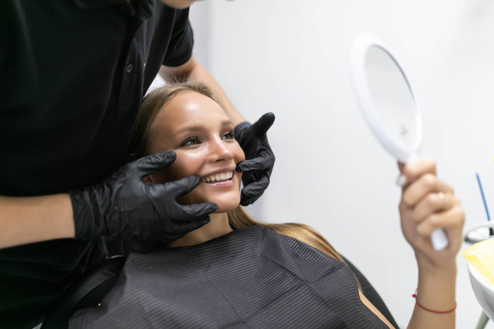 Patient Examining Smile in Mirror - Dental Crowns in Bakersfield Smiling woman at a dental clinic checking her teeth in a mirror, assisted by a professional. Perfect for cosmetic dentistry, smile makeovers, and aesthetic care. - Dental Crowns in Bakersfield