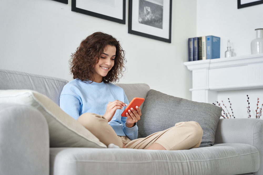 Young woman smiling while browsing her phone in a cozy living room. Great for topics like digital life, remote work, comfort, and home living. - Dentist in River Oaks