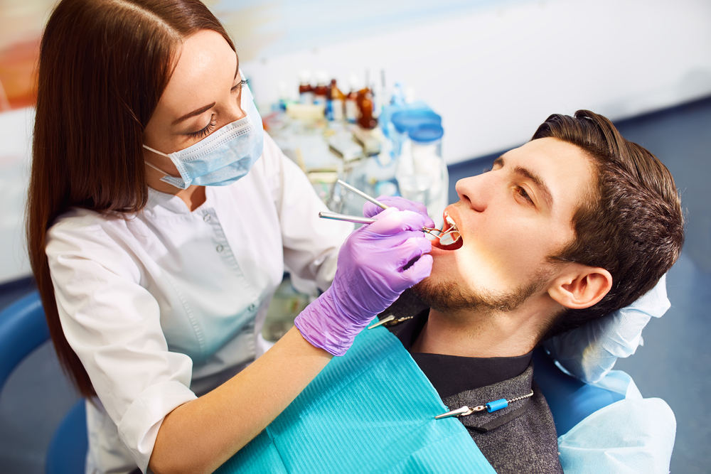 Dentist Performing Examination on Male Patient – Cost of Root Canal A dentist wearing a mask and gloves examines a male patient's mouth with dental tools, as he reclines in a chair with a blue bib – Cost of Root Canal.