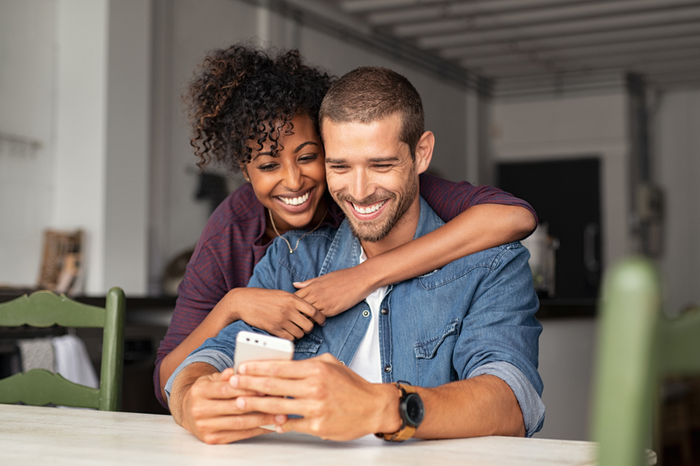 Joyful couple embracing while looking at a smartphone together in a cozy kitchen. Ideal for themes of connection, love, and modern lifestyle. - Dentist in River Oaks
