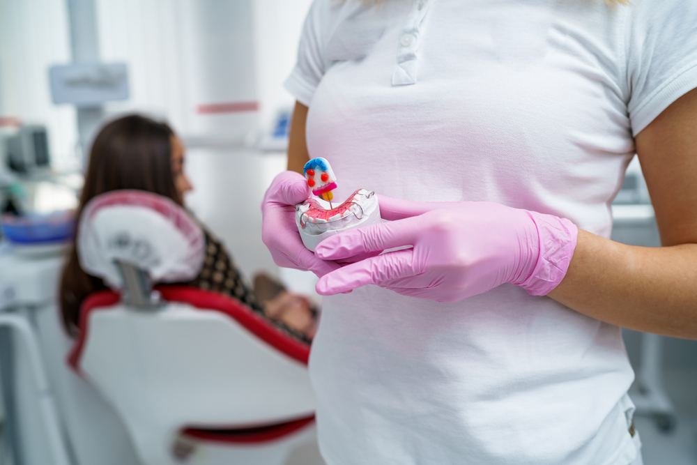 Dental Assistant Holding Tooth Model – Dentures Bakersfield A dental assistant wearing pink gloves holds a dental model with dentures and a color-coded tooth structure, while a patient sits in the background – Dentures Bakersfield