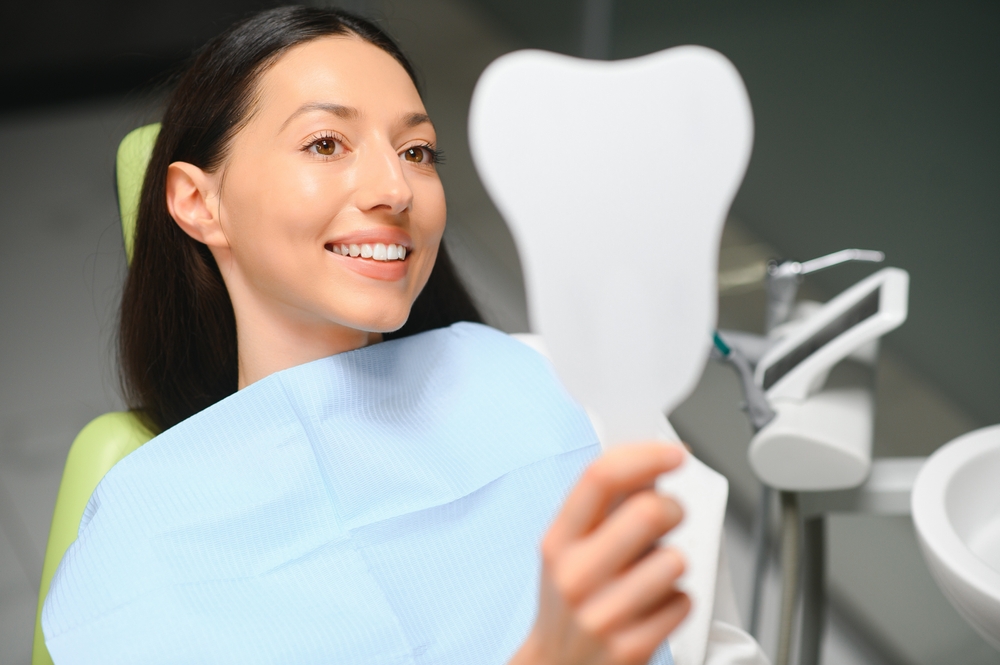 Woman Smiling in Dental Chair Holding Mirror – Alternative Root Canal Treatments A woman in a dental chair smiles while holding a tooth-shaped mirror, wearing a blue dental bib in a clinic setting – Alternative Root Canal Treatments