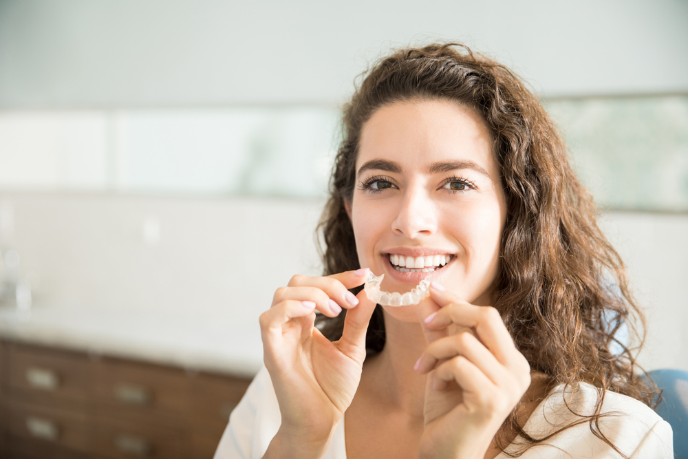 Smiling Woman Holding Clear Aligner – Braces vs Invisalign A woman with curly hair smiles while holding a clear aligner in a dental office, representing a modern alternative to traditional metal braces – Braces vs Invisalign