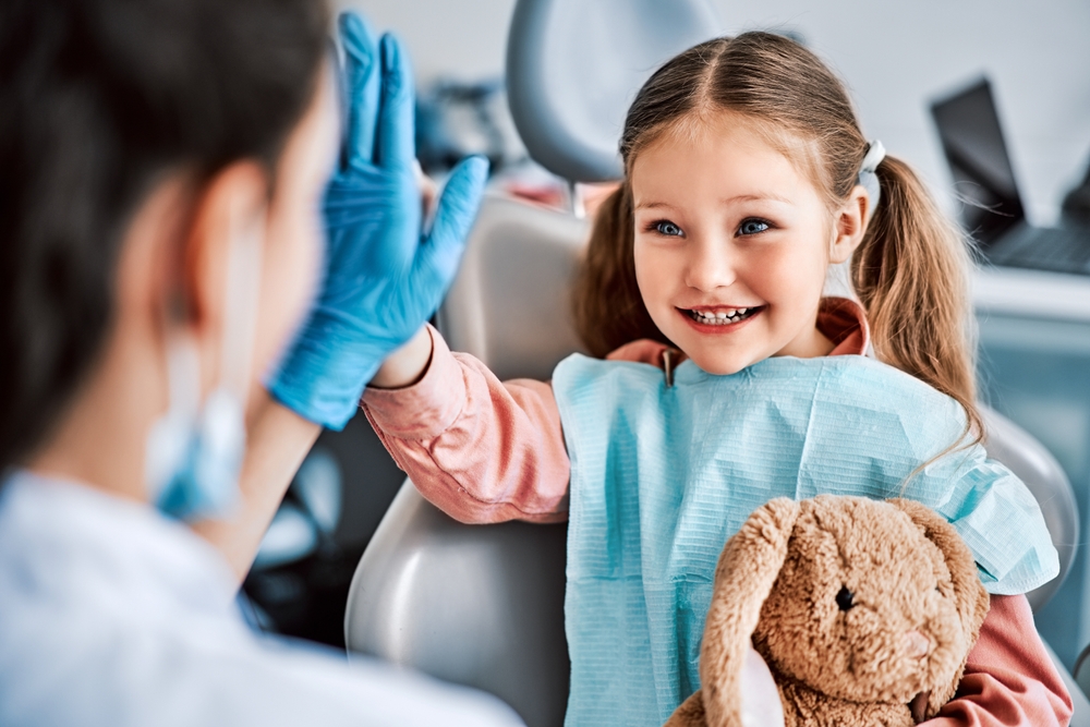 Smiling Girl at Dentist - Pediatric Dentist Bakersfield Happy young girl giving a high-five to the dentist while holding a stuffed toy. Perfect for promoting friendly, kid-focused dental care and trust-building. - Pediatric Dentist Bakersfield