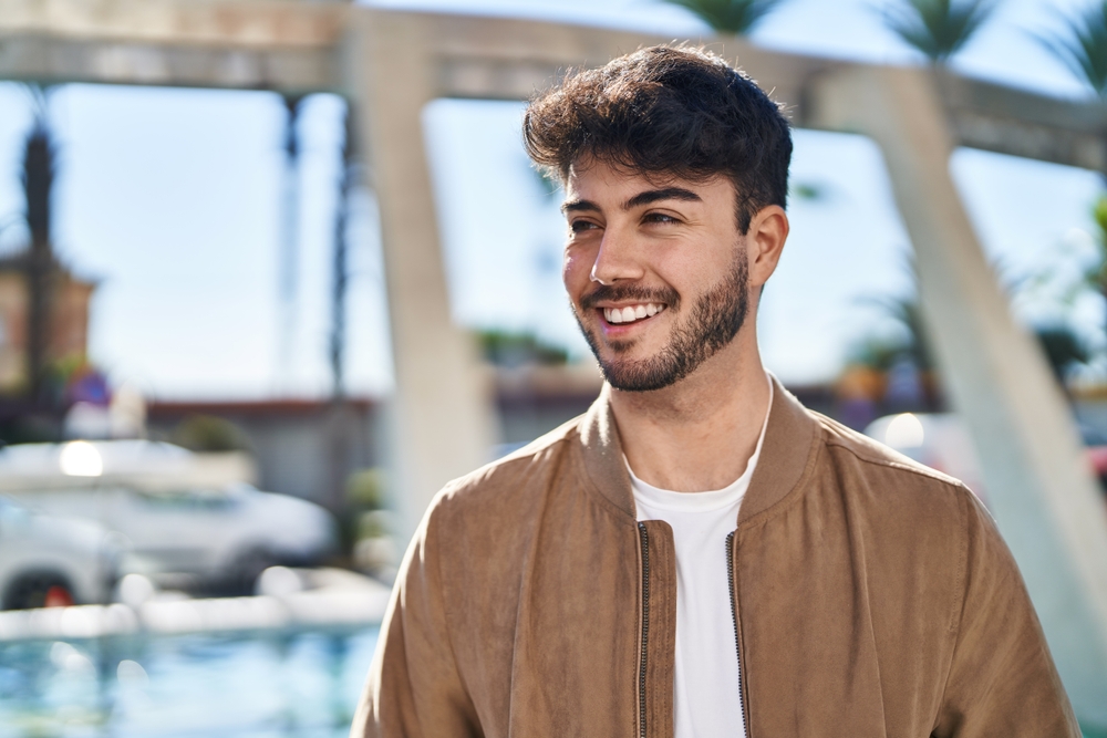 Smiling Young Man Outdoors - Cosmetic Dentist Cheerful man in a tan jacket smiling under bright sunlight. Great for lifestyle, fashion, and positive mood themes. - Cosmetic Dentist