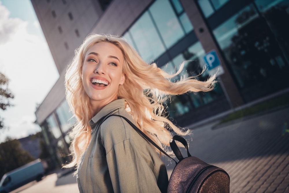 Joyful Blonde Woman Outdoors - Cosmetic Dentist Blonde woman laughing while walking outside with a backpack. Ideal for travel, freedom, youth lifestyle, and happiness content. - Cosmetic Dentist
