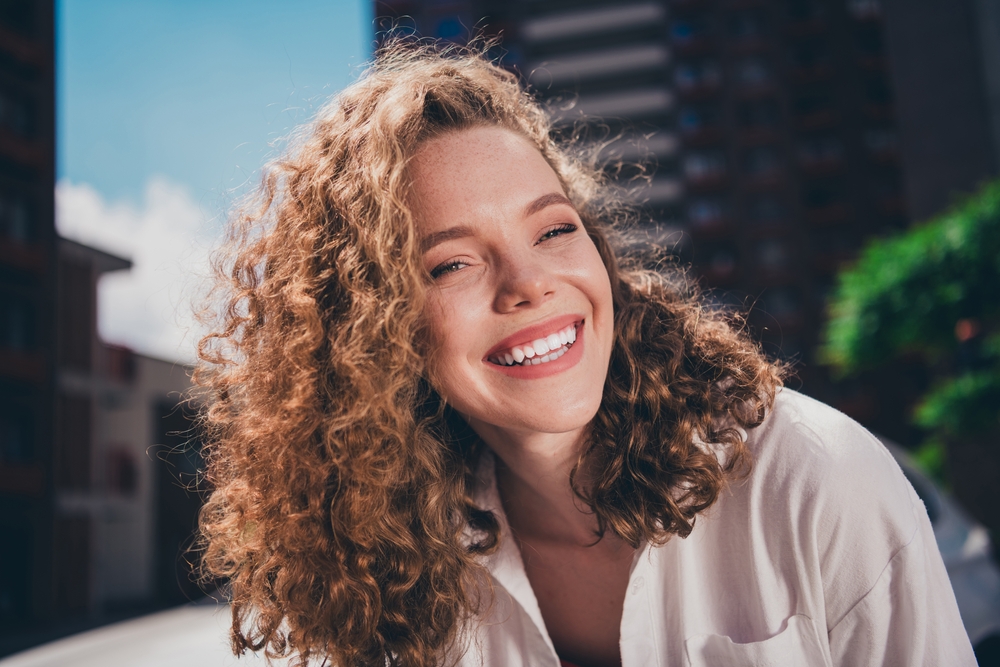 Joyful Woman Smiling Outdoors – Natural Beauty & Confidence - Dental Implants in Bakersfield Carefree woman with curly hair smiling brightly in natural light. Perfect for lifestyle, wellness, and self-confidence content. - Dental Implants in Bakersfield