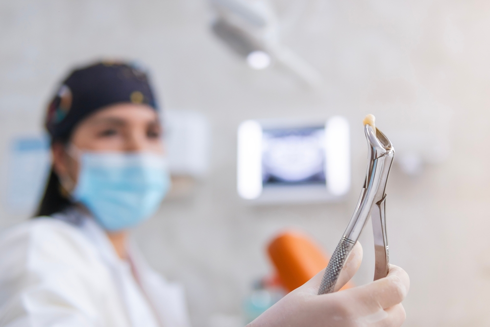 A masked dentist holds up a dental forceps gripping a recently extracted tooth, with the blurred patient and X-ray screen in the background – Tooth Extraction Costs