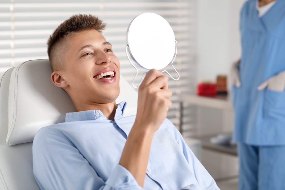 Young Man Smiling in Mirror After Dental Treatment – Veneers in Bakersfield A young man in a light blue shirt smiles while looking into a handheld mirror at the dentist’s office, appearing pleased with his teeth – Veneers in Bakersfield